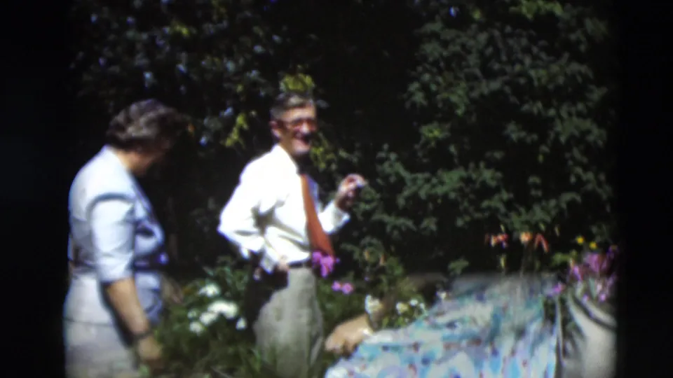 A woman in a light dress bends near garden flowers as a man in a white shirt and tie smiles in the 1951 Paducah home-movie scene.