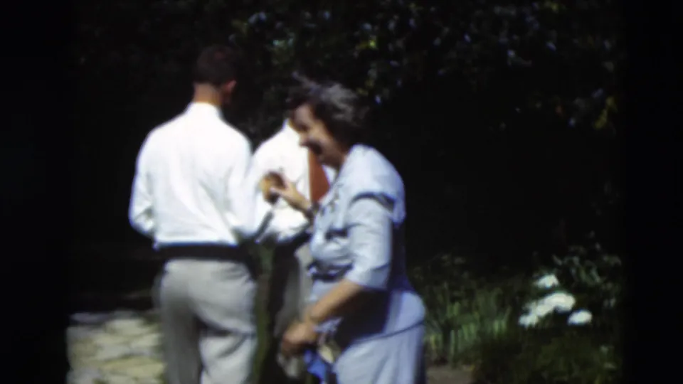 Adults in light mid-century clothing move through a Paducah garden, with a stone path, flowers, and dark foliage visible in the frame.