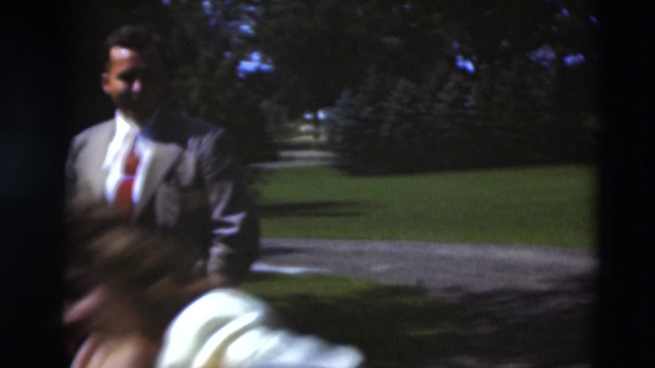Man in a suit stands on a lawn while a blurred figure crosses the foreground.