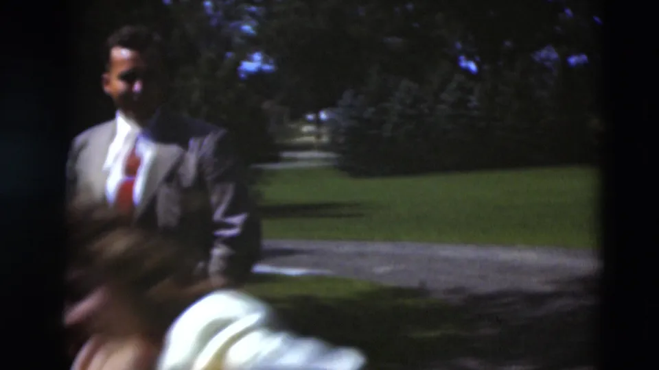 A suited man stands on a sunlit lawn in the Paducah garden scene as a blurred woman passes close to the camera.