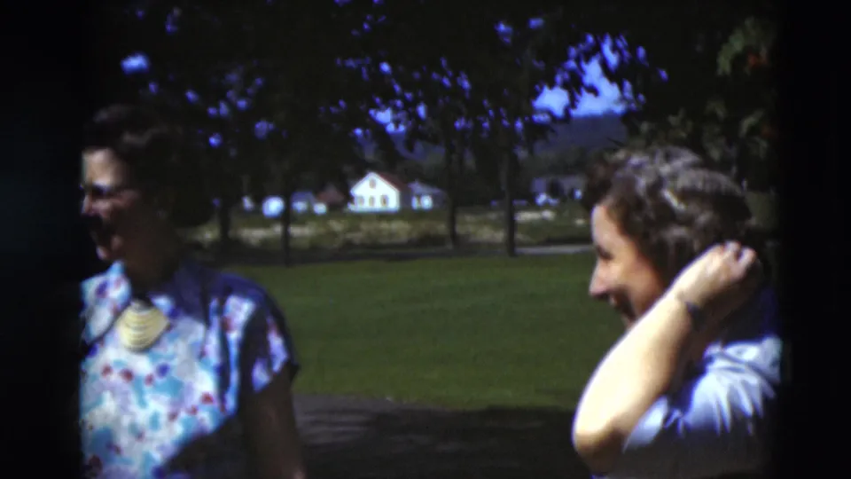 Two women in patterned and light clothing appear to laugh or talk on a Paducah lawn, with trees and a distant house visible.