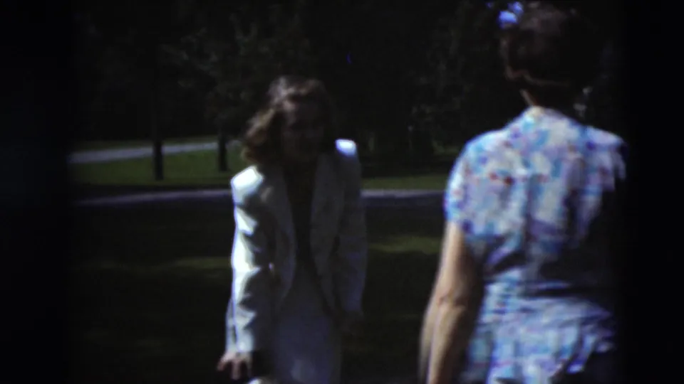 Two women in light and patterned dresses move through the shaded Paducah garden area near a road or driveway.