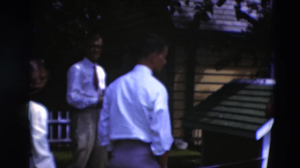 1951 Paducah home-movie frame with adults standing in shade near a wooden yard structure and fence.