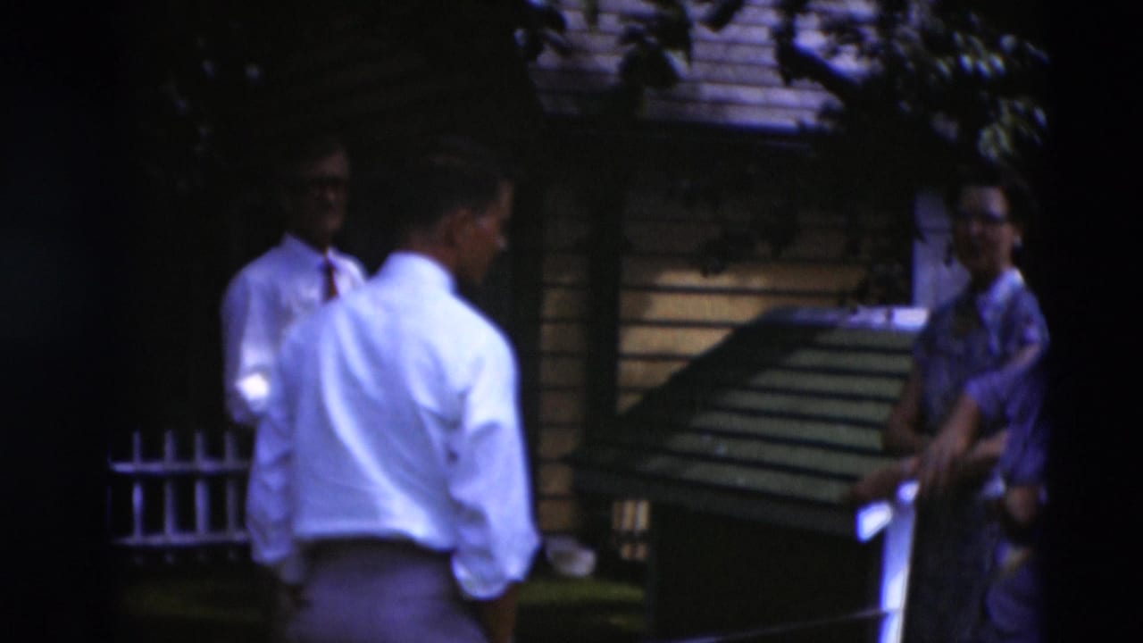 1951 Paducah home-movie frame showing adults conversing in a shaded backyard near a small wooden structure and house siding.