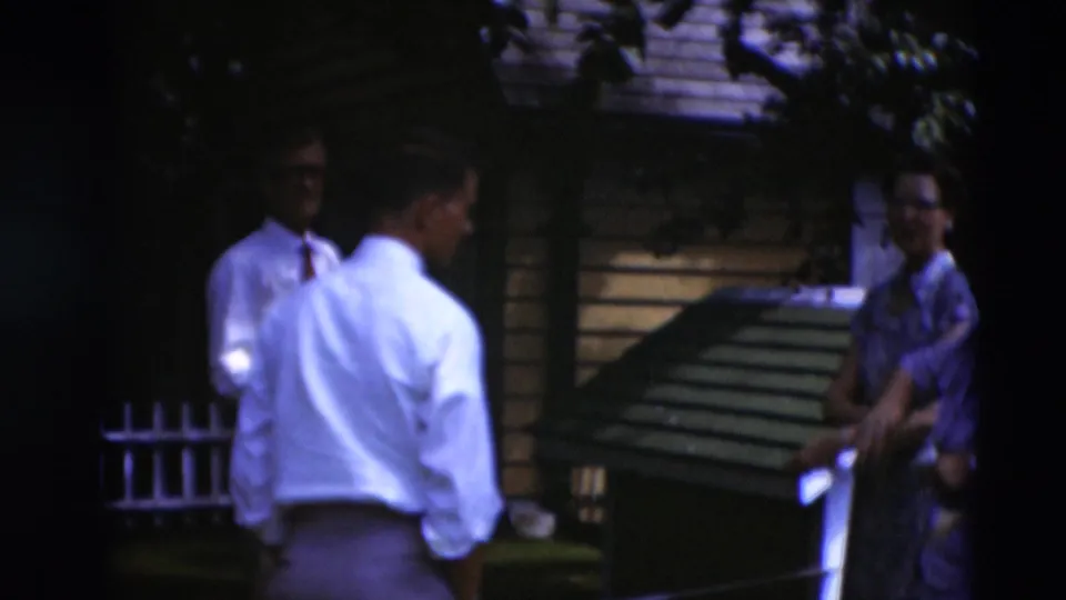 1951 Paducah home-movie frame showing adults conversing in a shaded backyard near a small wooden structure and house siding.