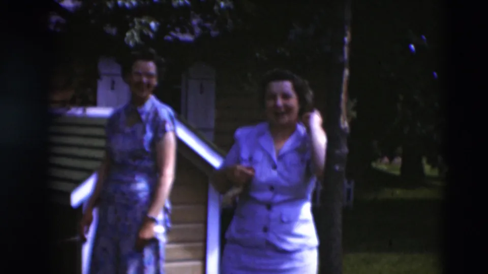 1951 Paducah frame of two women in dress clothing moving casually near a white railing and small backyard structure.