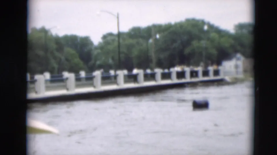 Paducah flood footage showing muddy water beside a bridge or levee wall, trees, and a floating barrel-like object.