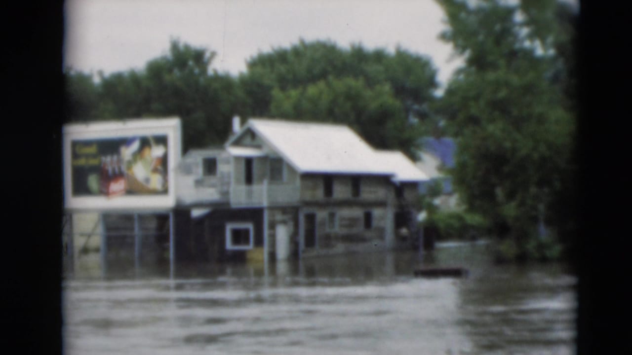 Floodwater surrounds houses and a roadside billboard in a tree-lined neighborhood.