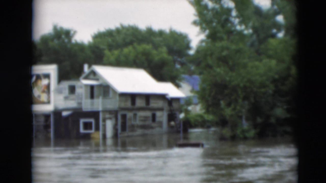 Paducah floodwater surrounds residential buildings, trees, and a partially readable billboard in this 1951 8mm frame.