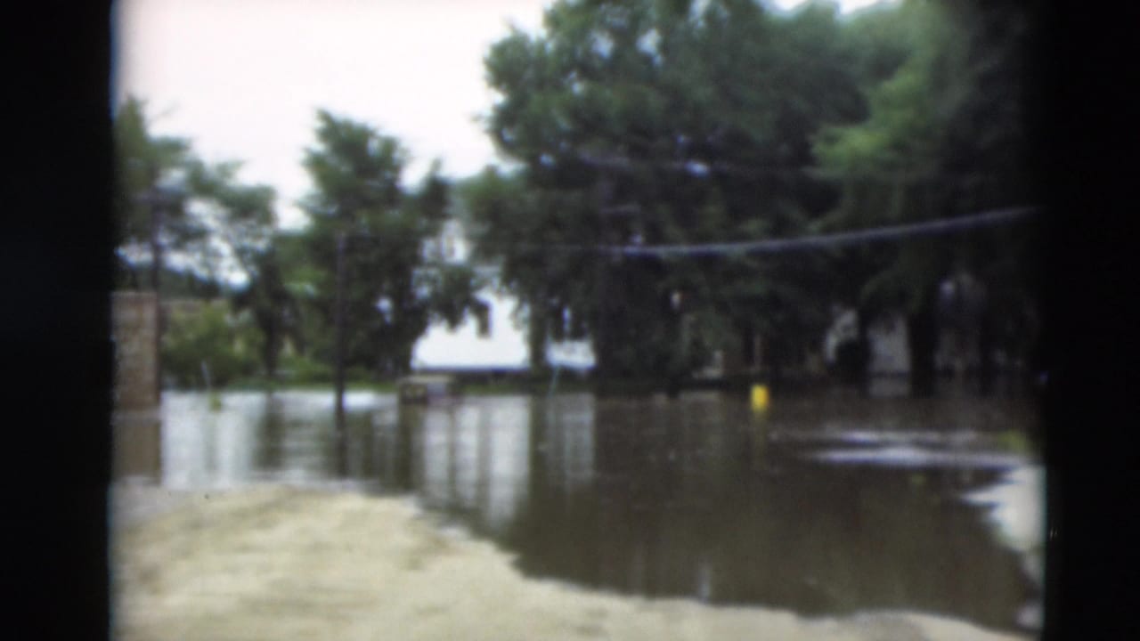 Floodwater fills a neighborhood street beside trees and small buildings under a cloudy sky.