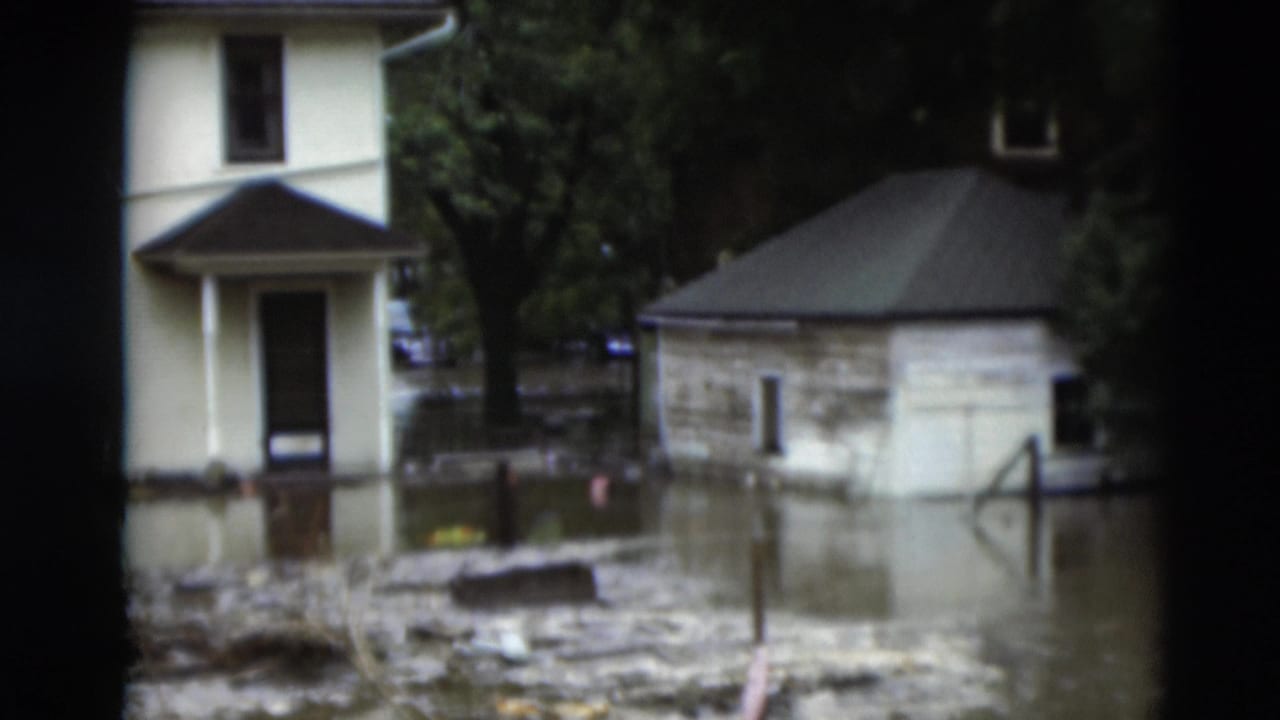 Muddy floodwater surrounds a house, trees, and a small stone building.