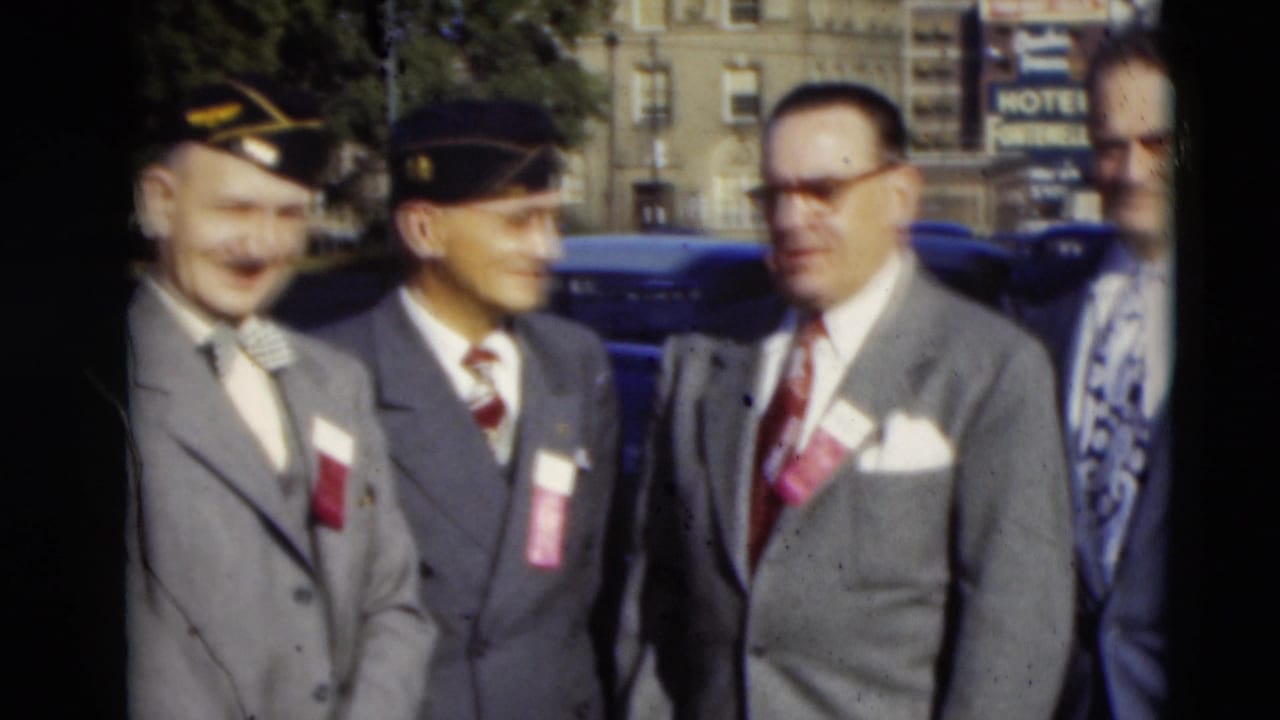 Four suited men with badges stand on a city street beside parked cars.