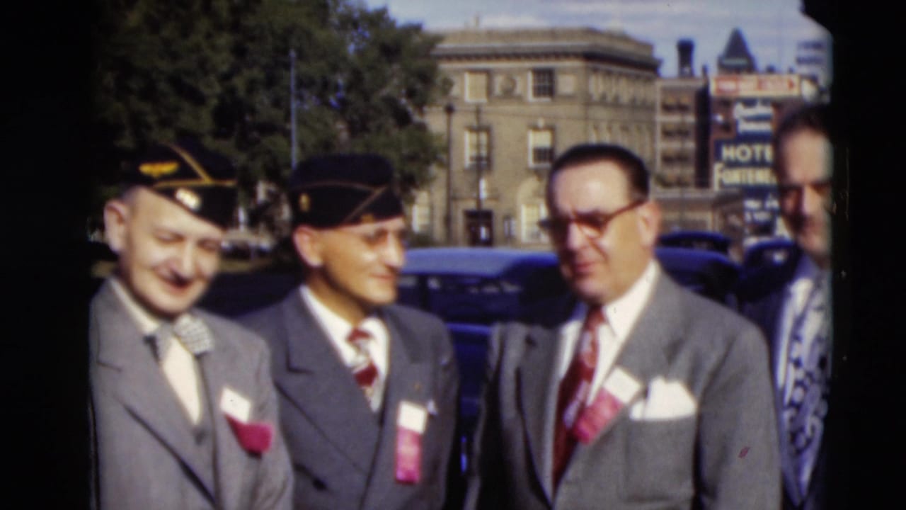 Three suited older men in caps pose outdoors beside parked cars and downtown buildings.
