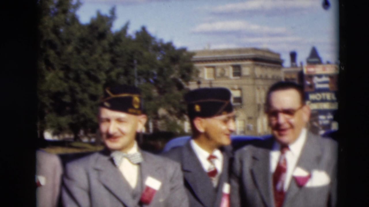 Three older men in suits and caps stand close together with a hotel sign in the background.
