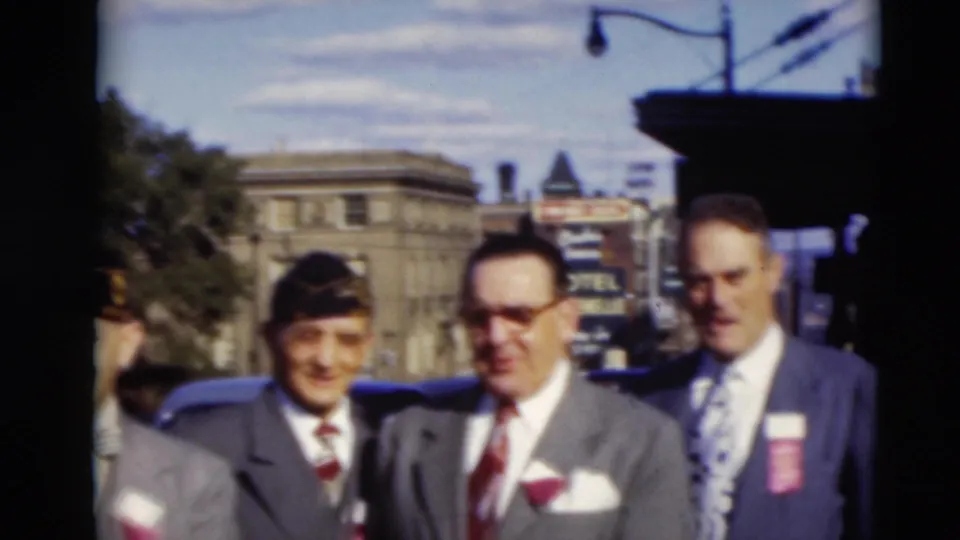 Several suited men, some with caps and red boutonnieres, stand in a Paducah street scene partly cropped by handheld 8mm framing.