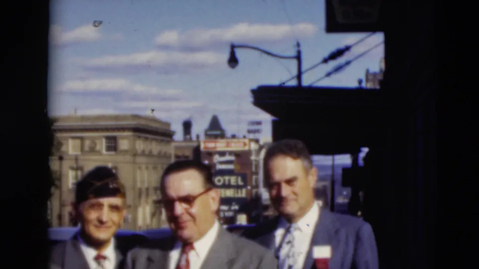 Three adult men in suits, one wearing a dark cap, stand in a 1951 Paducah downtown scene with a hotel sign, streetlamp, and building in the background.