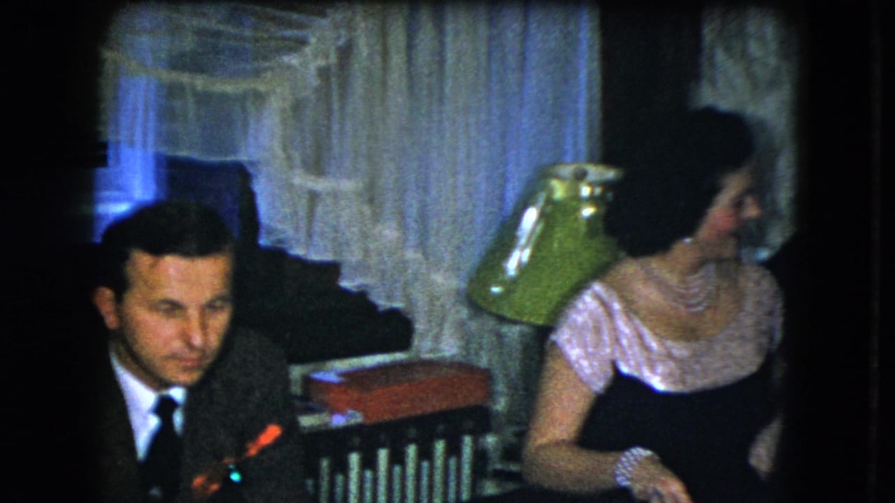 1951 Cleveland home-movie frame with a suited man and a woman in pearls seated near curtains and a green lamp.