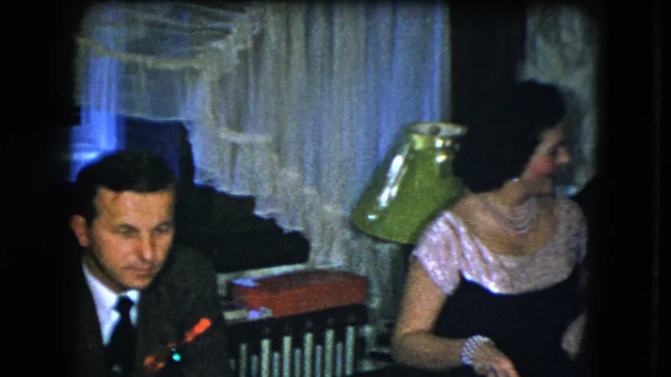 1951 Cleveland home-movie frame with a suited man and a woman in pearls seated near curtains and a green lamp.
