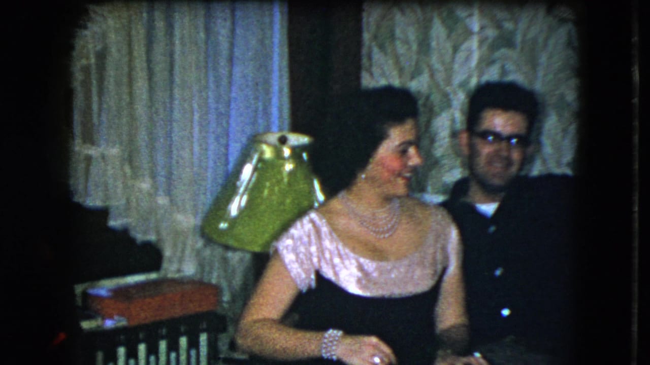 Woman in a dress turns toward a man with glasses near a green lampshade.