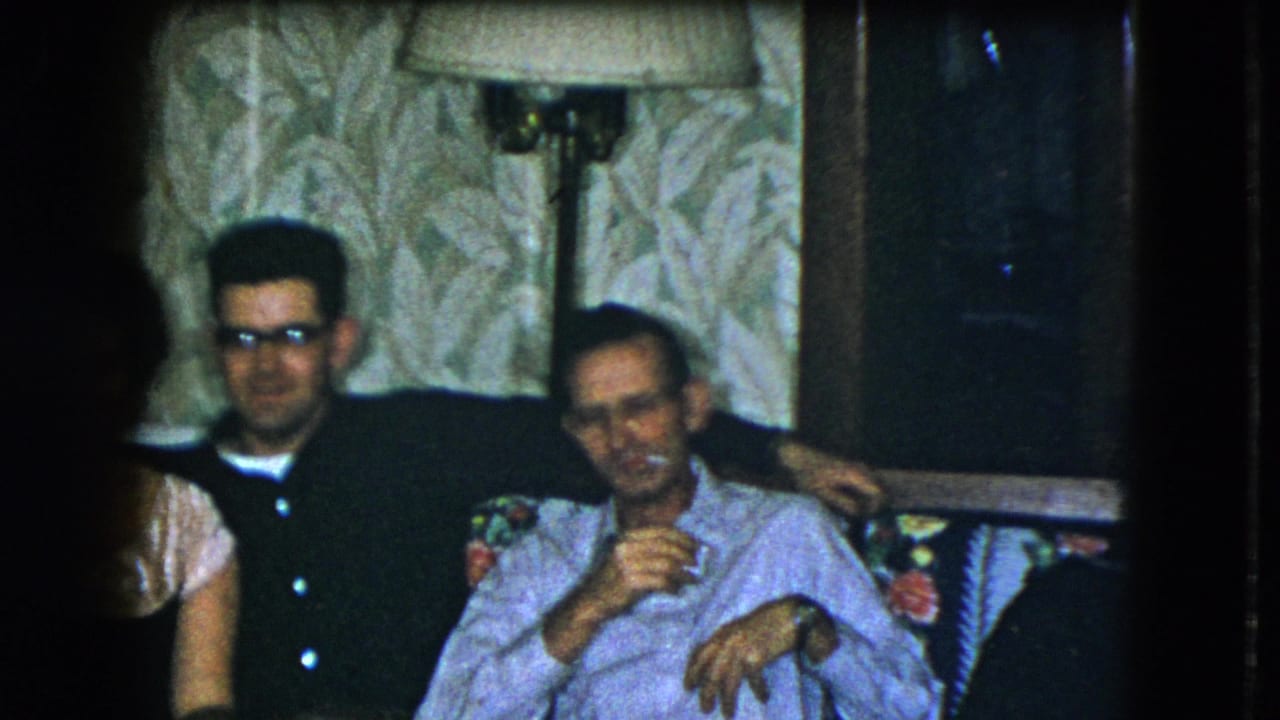 Two adult men remain seated in the Cleveland living room, with a partial figure at left and one man smoking casually.