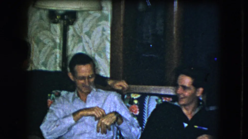 The home-movie frame shows a man in a light shirt beside a smiling man in a dark shirt in the same 1951 Cleveland living room.