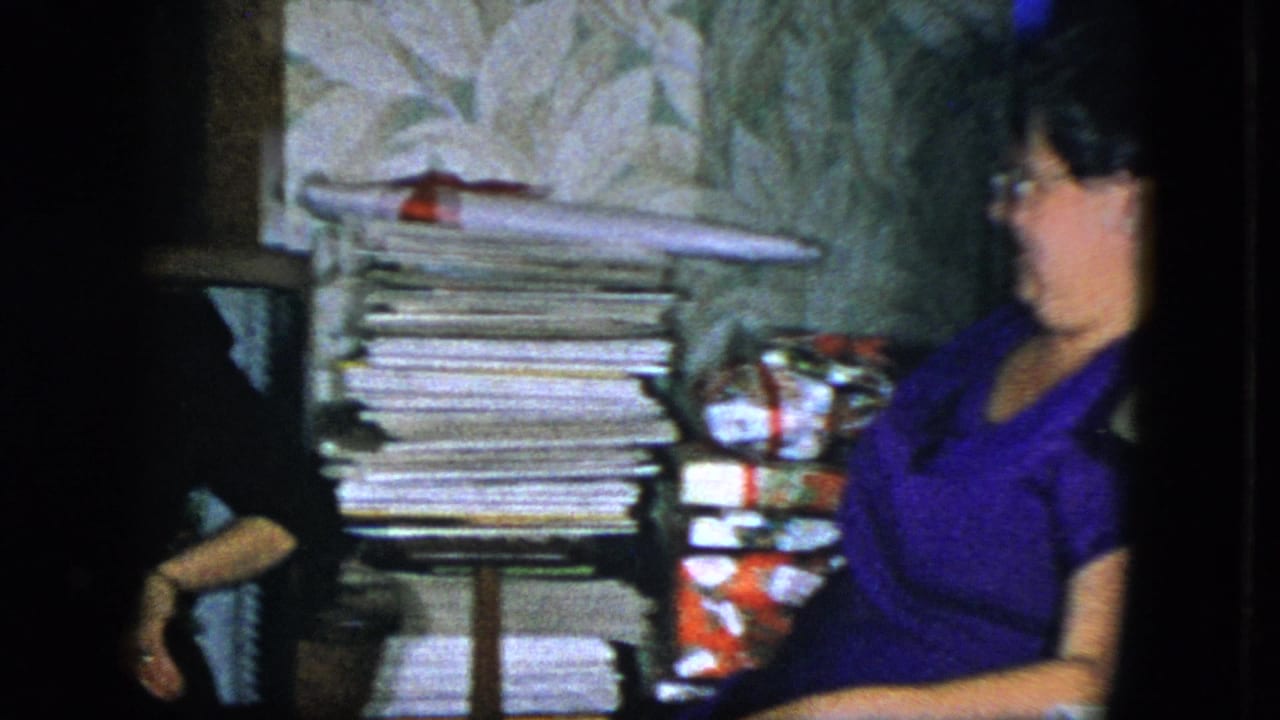 In a 1951 Cleveland home-movie frame, a seated woman in glasses and a purple dress sits beside magazines and wrapped Christmas gifts.