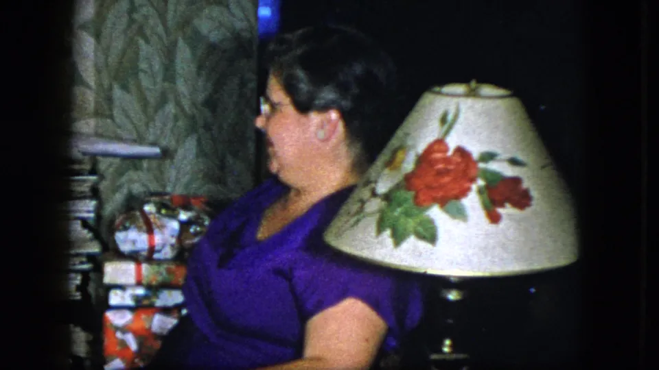 The woman turns in profile beside a floral lampshade, patterned wallpaper, magazines, and wrapped gifts in the Cleveland interior.