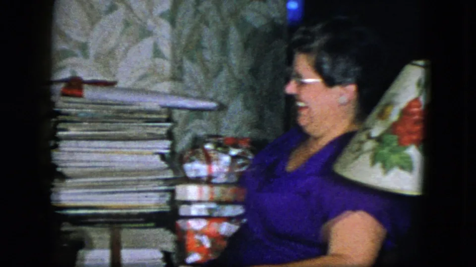 The seated woman smiles or laughs beside stacked magazines, wrapped gifts, wallpaper, and a floral lampshade in a Cleveland home setting.