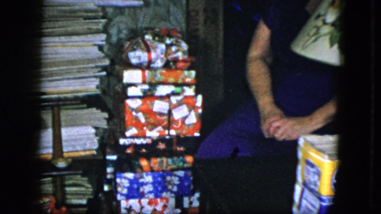1951 Cleveland home-movie frame showing a woman seated beside stacked wrapped Christmas gifts in a private interior.