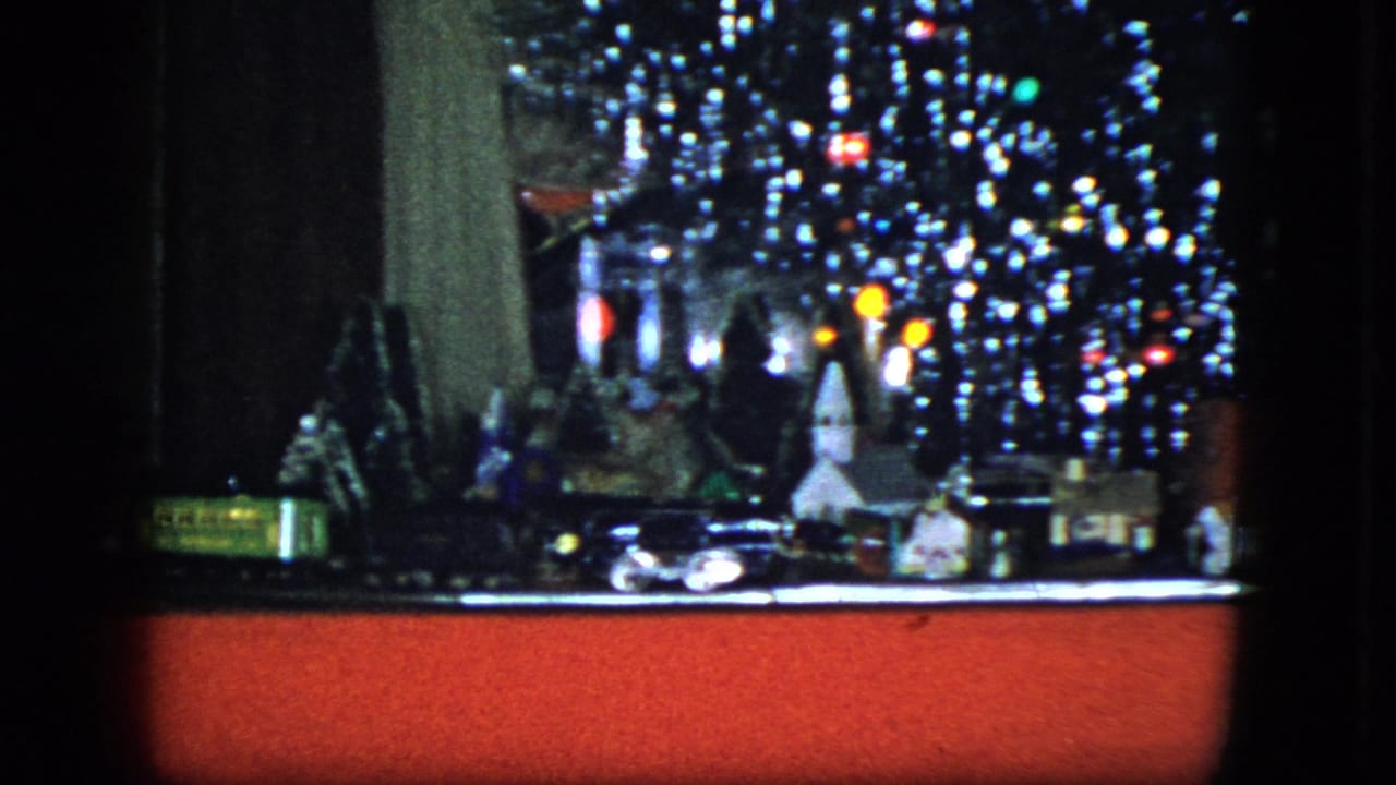 1951 Cleveland home-movie frame with a model train passing miniature buildings beneath a tinsel-covered Christmas tree and red display base.