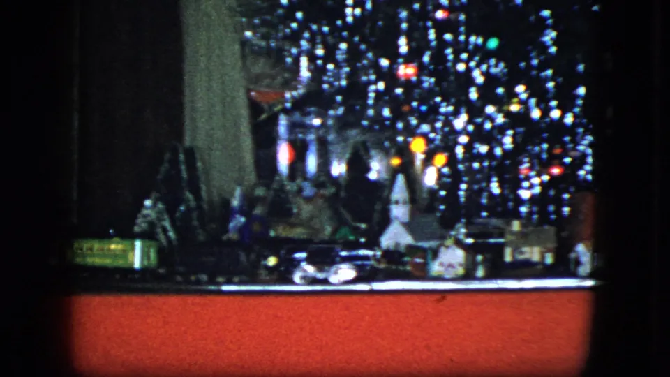 1951 Cleveland home-movie frame with a model train passing miniature buildings beneath a tinsel-covered Christmas tree and red display base.