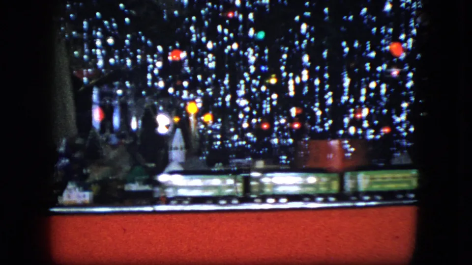 1951 Cleveland 8mm frame showing model train cars moving across the front of a miniature Christmas village under sparkling tinsel lights.