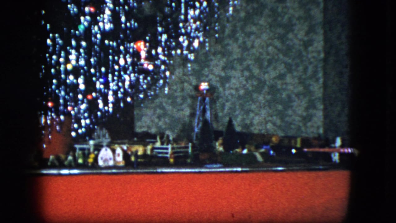 1951 Cleveland home-movie frame showing a model train layout beside a lit Christmas tree on a red platform.
