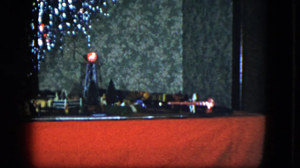 1951 home-movie still showing a miniature town and rail layout under a bright red Christmas tree light.