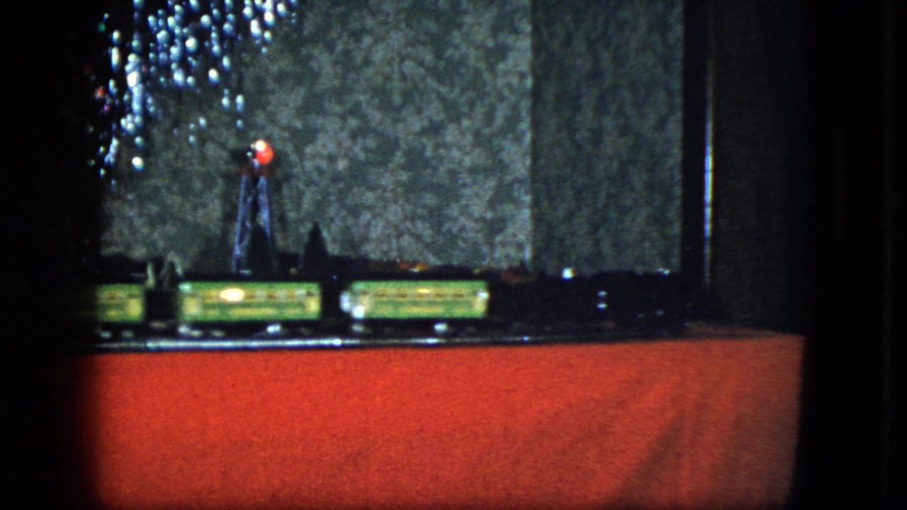 Model train layout on a red foreground with tree lights at left and a small tower near the center.