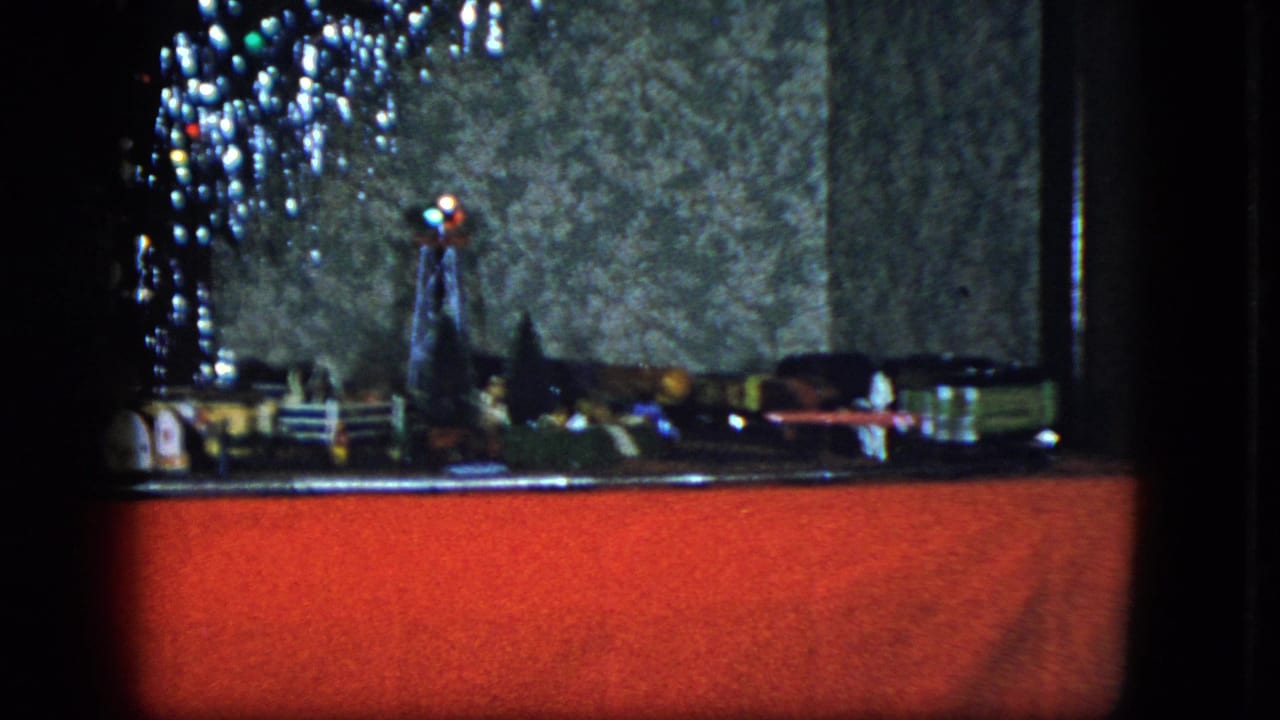 Miniature train layout with a red foreground, small buildings, and Christmas lights along the left edge.