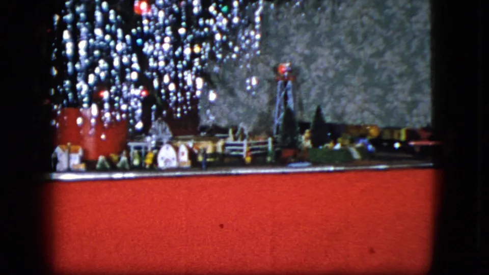 1951 Cleveland home-movie frame showing a miniature holiday railroad town with tiny houses, tinsel decorations, a dark backdrop, and a red display base.