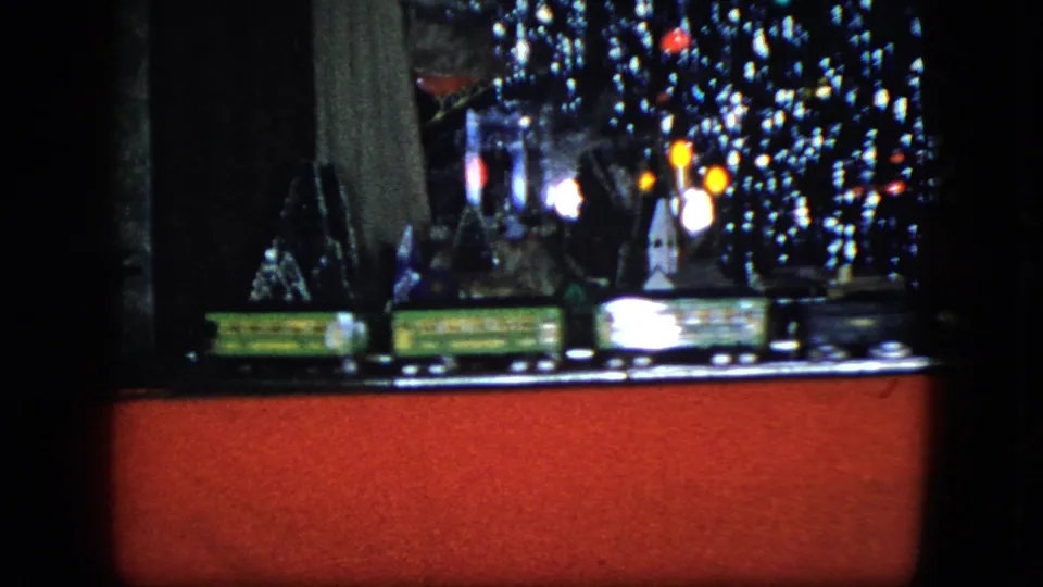 1951 Cleveland 8mm frame showing a toy railroad village with glowing holiday lights, tinsel, small buildings, and a dark indoor background.