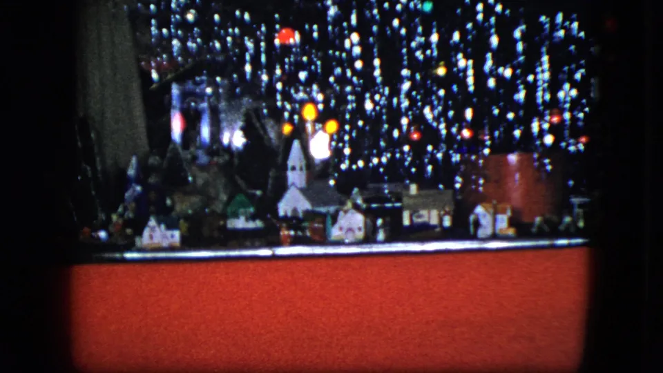 1951 Cleveland home-movie frame showing a compact toy train village scene with tinsel, glowing bulbs, small houses, and a red display base in an indoor setting.