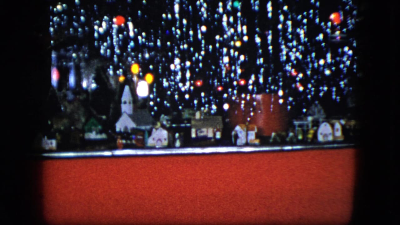 1951 Cleveland home-movie frame of a Christmas model train setup with a miniature church, houses, red train car, and lights.
