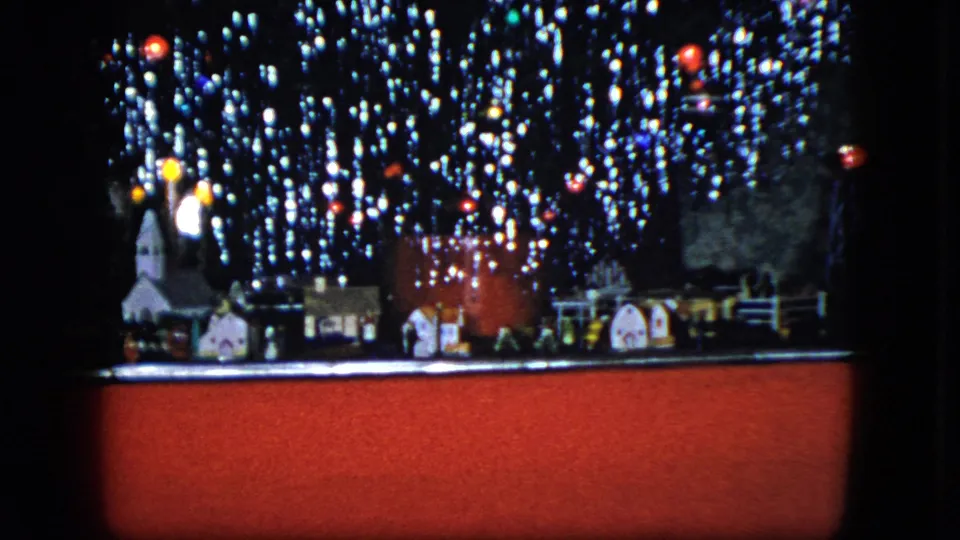 1951 Cleveland home-movie frame of a Christmas model train scene with miniature buildings, lights, and a red foreground edge.