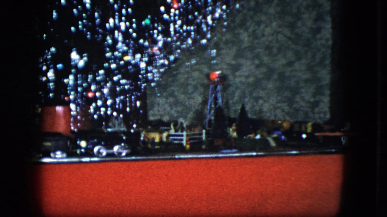 Model train Christmas display with red fabric base, glowing signal, miniature buildings, and tree lights.