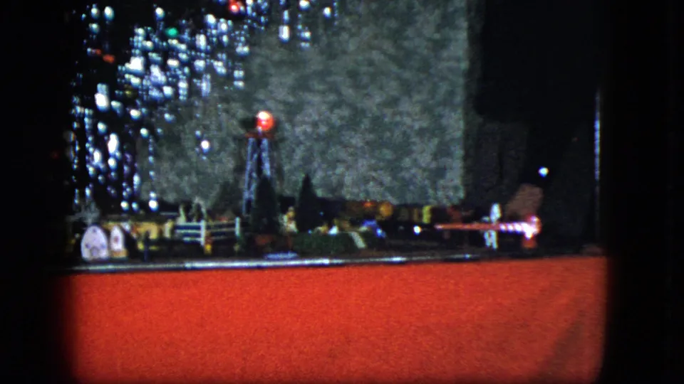 Cleveland 1951 home-movie still showing a Christmas model train layout on red fabric, with miniature buildings, tree lights, and a nearby hand.