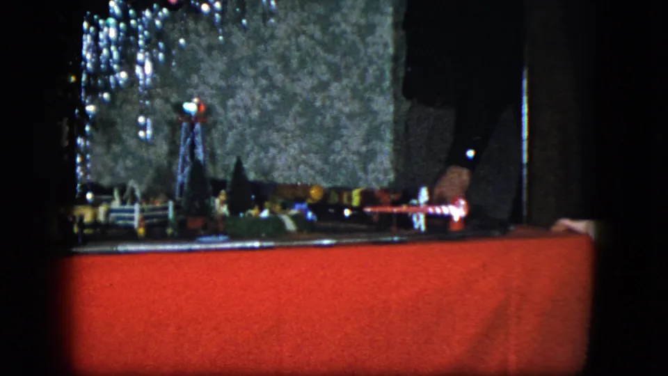 1951 Cleveland 8mm frame of a Christmas model railroad display with a red base, illuminated signal, miniature objects, and a hand at the right.