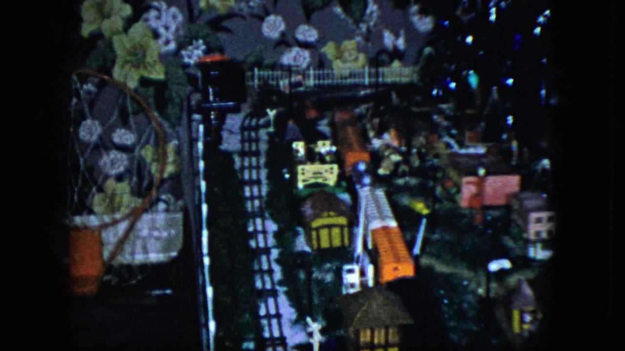 1951 Cleveland home-movie frame showing a tabletop model train display with tracks, small structures, and floral wallpaper behind it.