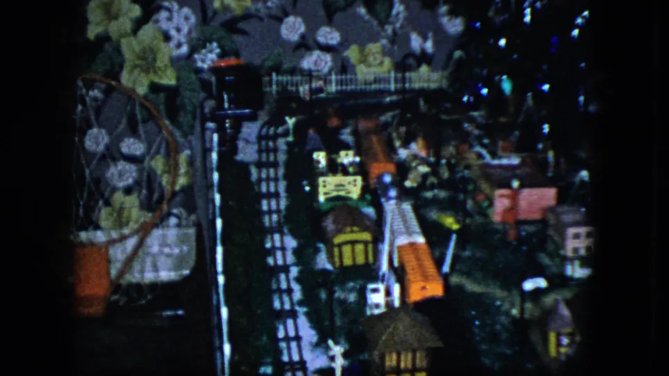1951 Cleveland home-movie frame showing a tabletop model train display with tracks, small structures, and floral wallpaper behind it.