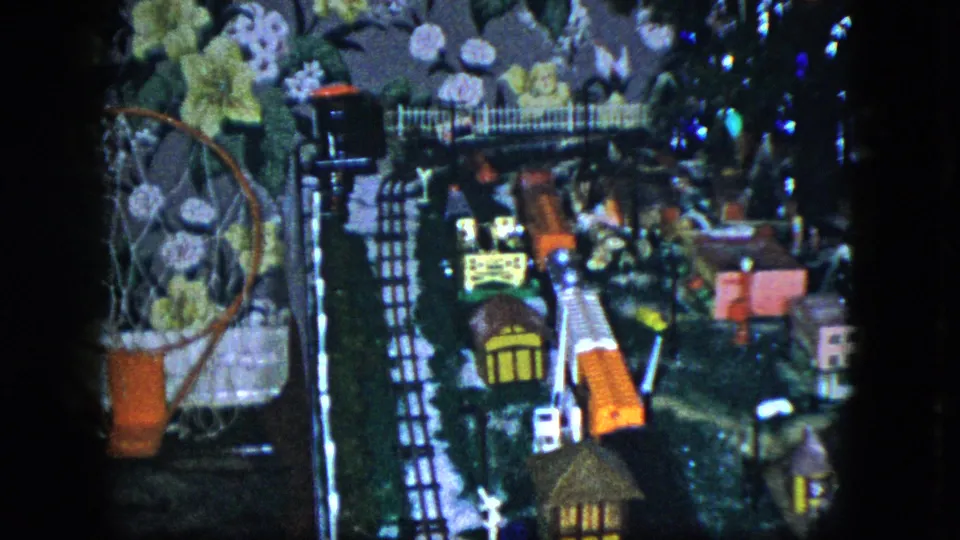 1951 Cleveland home-movie still showing the model train set from above, with tiny buildings, track, and a patterned wall behind it.