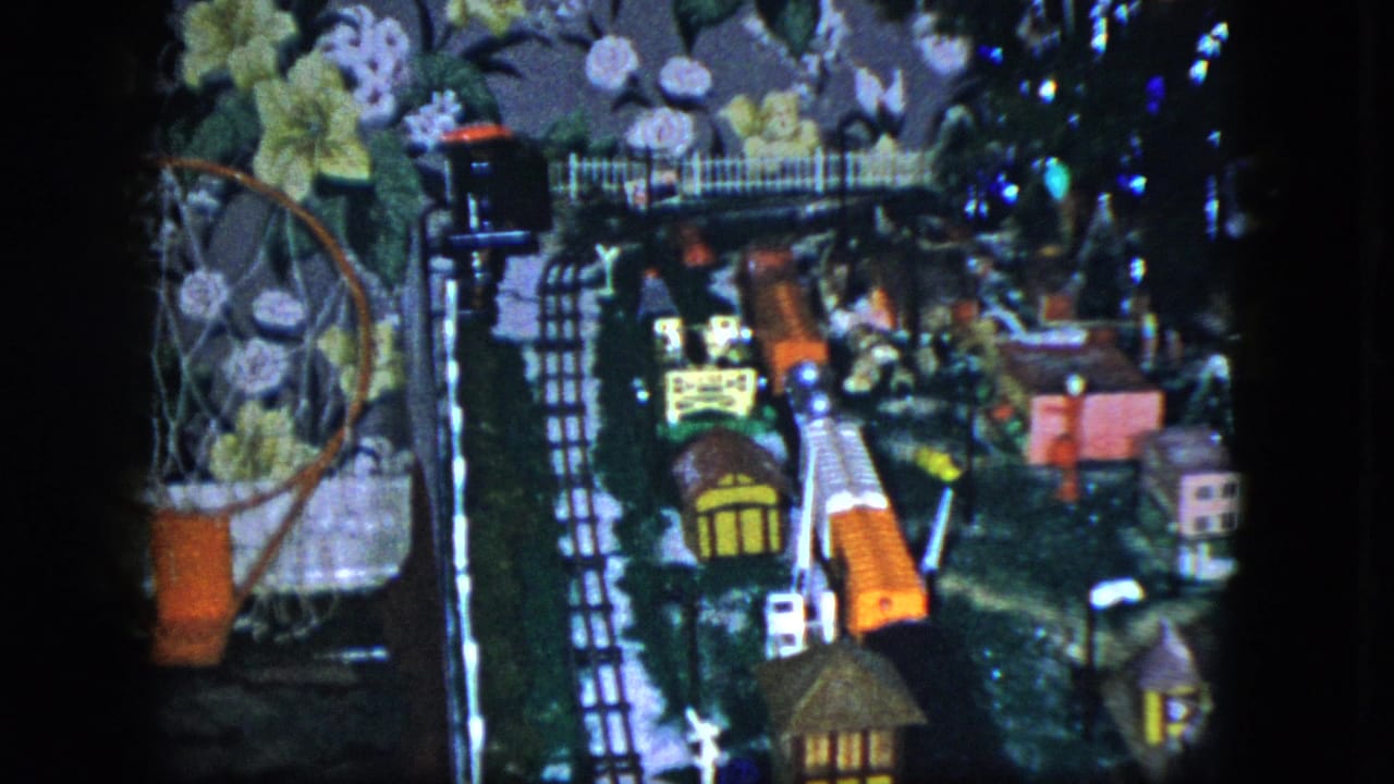 1951 Cleveland home-movie frame showing a colorful miniature train layout with tiny houses, fence, bridge, and floral wallpaper in the background.
