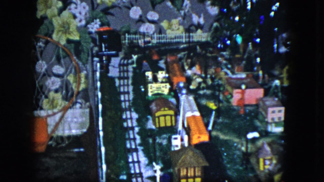 1951 Cleveland frame of a miniature railroad display with orange train cars, small buildings, a white fence, and floral wallpaper behind the layout.