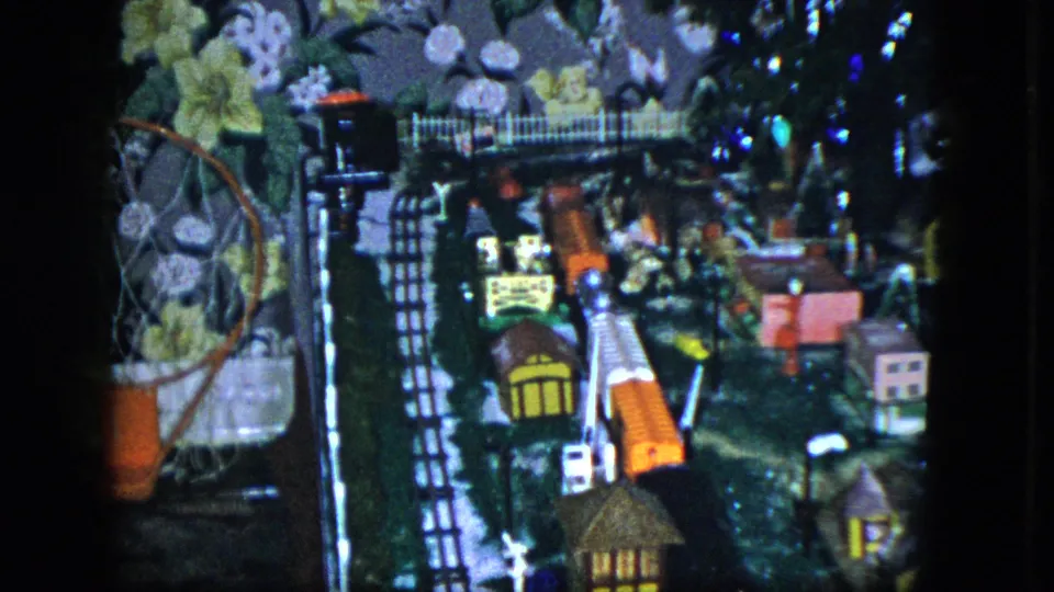 1951 Cleveland home-movie frame showing the model train village, with track, small houses, a fence, bridge, floral wallpaper, and a basketball hoop at left.